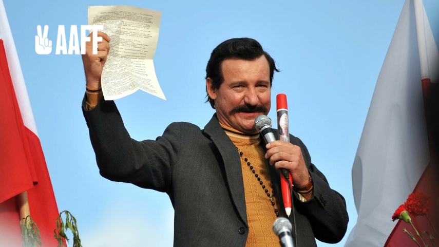 Robert Więckiewicz - Mężczyzna stojący na scenie trzyma w górze kartkę z tekstem i mówi do mikrofonu, a po bokach widoczne są flagi Polski.